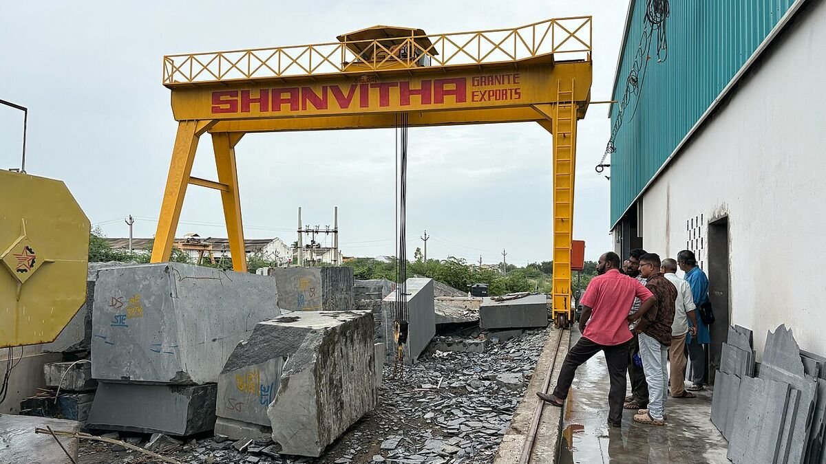 Buyers inspecting large granite blocks beneath a yellow gantry crane at a granite export yard